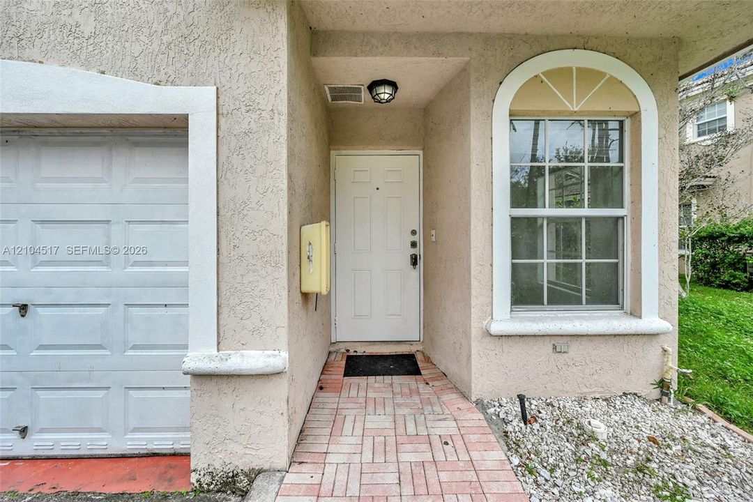 Stucco exterior entry featuring a brick-paver walkway, single-car garage with white panel door, arched window with decorative muntins, and a covered porch with a flush-mount light fixture