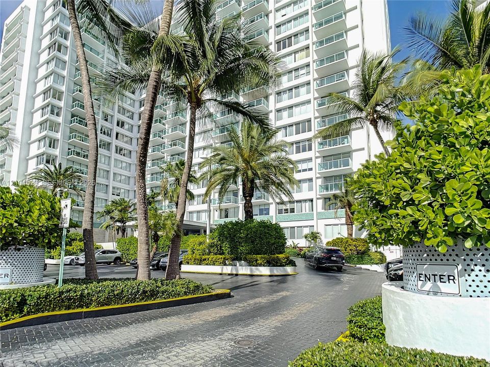 High-rise residential exterior featuring white concrete facade, stacked glass-railed balconies, paved circular motor court, and established tropical landscaping