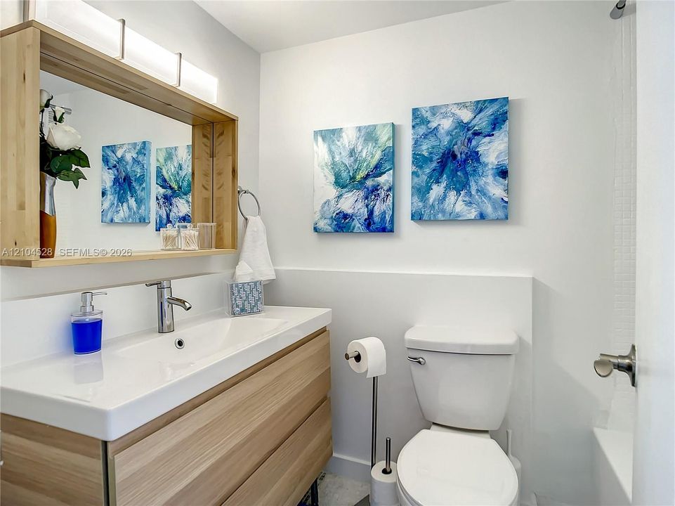Modern bathroom featuring a wood-finish vanity with a white rectangular countertop and single-handle chrome faucet, a white toilet with chrome supply line, white subway tile wainscoting, recessed ceiling lighting, a wood-framed mirror with integrated lighting, and blue abstract artwork