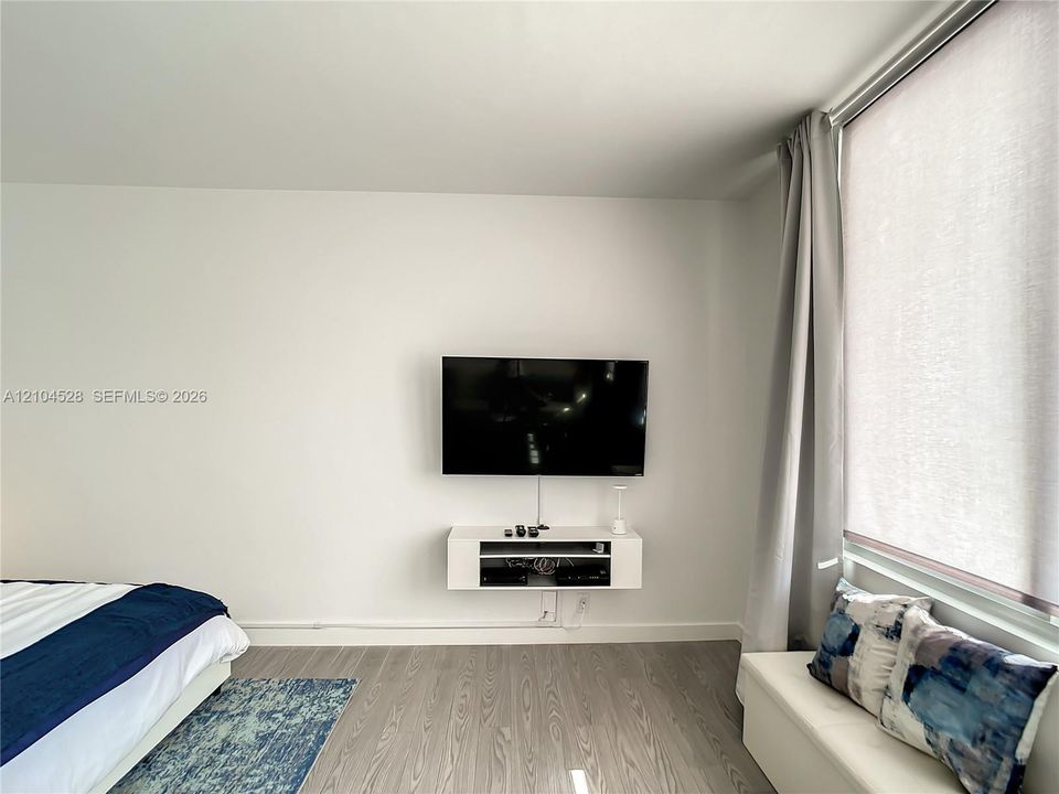 Neutral interior featuring wood-finish flooring, a wall-mounted floating media console, and a large window with roller shades