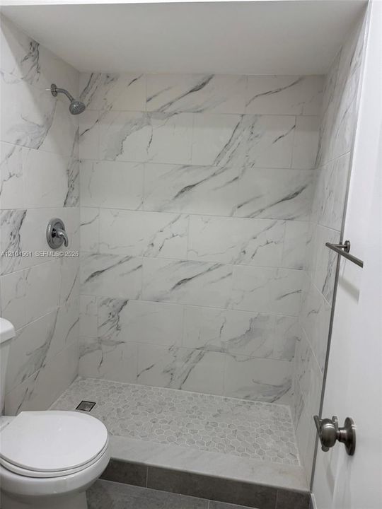 Walk-in shower featuring large-format white stone-finish tile surround, pebble-texture floor tiling, and chrome wall-mounted fixtures