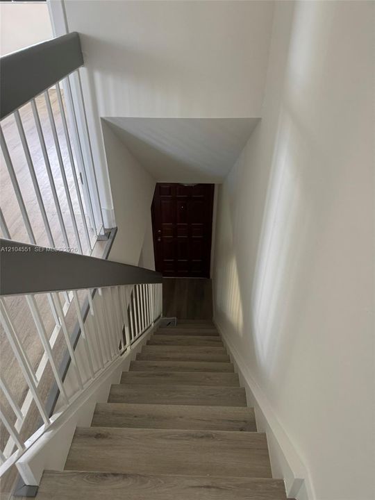 Staircase featuring wood-finish treads, a white metal balustrade, and a dark-toned handrail, ending at a multi-panel door