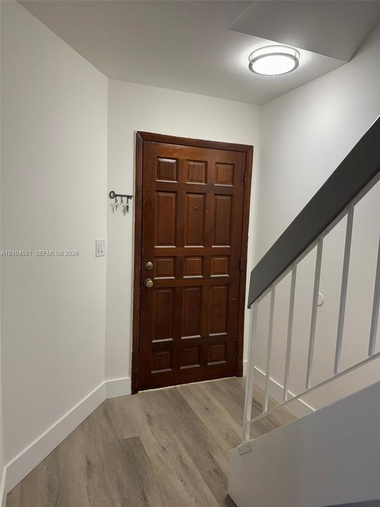 Entryway featuring a solid wood-finish door with multi-panel detailing, neutral wood-finish flooring, and a staircase with a white railing and dark-toned handrail, illuminated by a flush-mount circular ceiling fixture