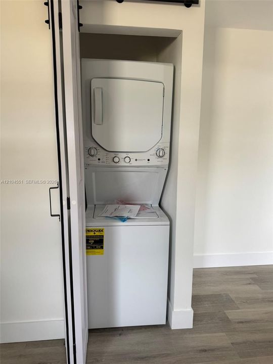 Stacked laundry unit enclosed by a white sliding barn door featuring matte black hardware and wood-finish flooring