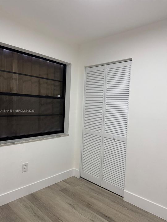 Room featuring wood-finish flooring, white louvered closet doors, and a window with a black frame and stone sill