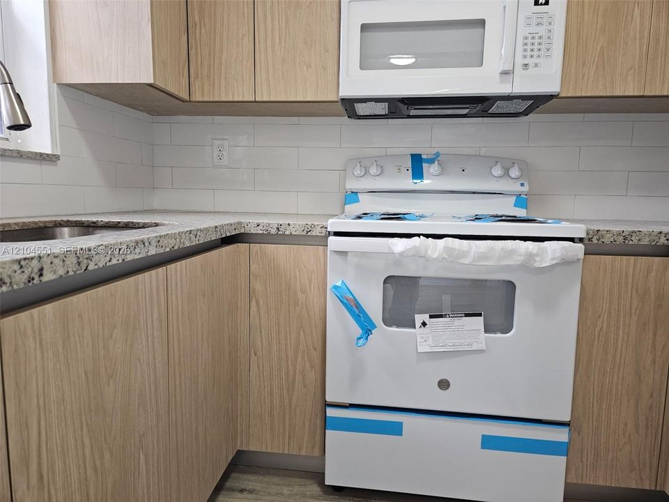 Kitchen featuring flat-panel wood-finish cabinetry, speckled stone-finish countertops, white subway tile backsplash, and white appliances