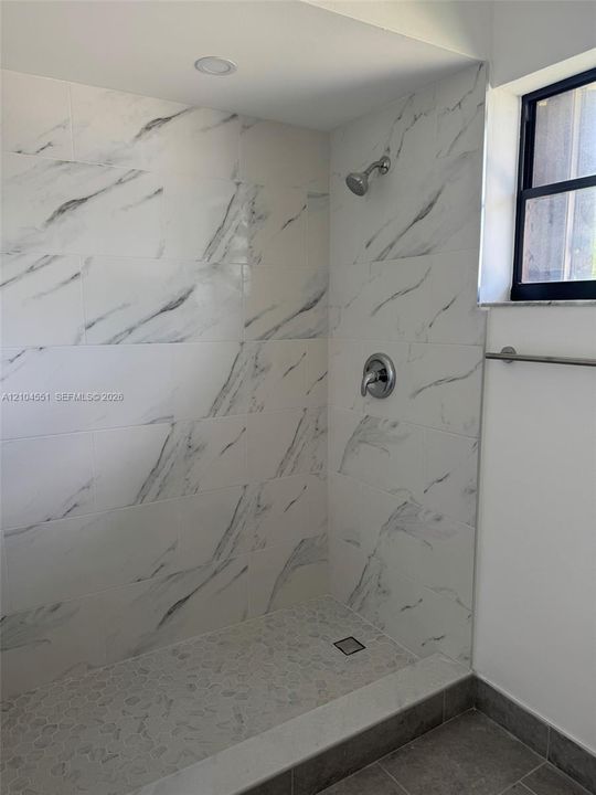 Walk-in shower featuring large-format stone-patterned wall tile, pebble-textured flooring, recessed lighting, and a metal-finish fixture