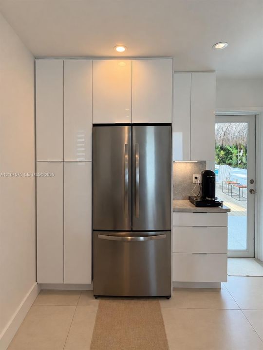 High-gloss white cabinetry, stainless steel French-door refrigerator, stone-surface countertop, recessed ceiling lighting, and large-format tile flooring