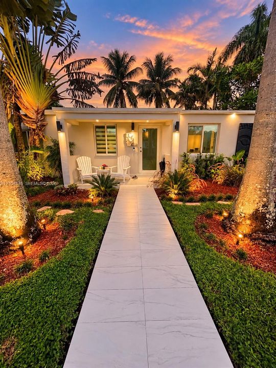 Covered front porch featuring large-format tile flooring, porcelain-tiled walkway, manicured landscaping, and mature palm trees