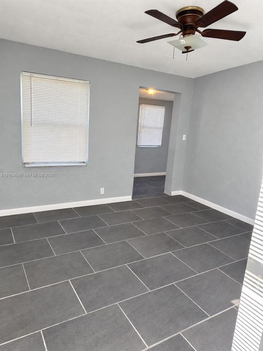 Room interior featuring large-format dark grey tile flooring, neutral wall paint, white horizontal blinds, and a multi-blade ceiling fan with integrated lighting