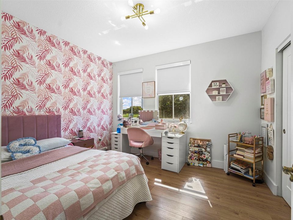 Bedroom featuring wood-finish flooring, dual windows with roller shades, a gold-tone multi-arm chandelier, and a decorative patterned accent wall