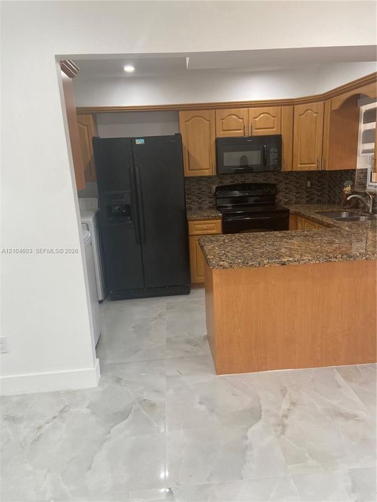 Wood-finish cabinetry, black appliances, tile backsplash, granite countertops, and polished tile flooring