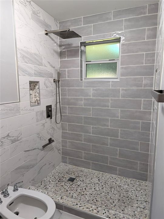 Walk-in shower featuring gray subway-style wall tile, contrasting white stone-finish wall accents, a pebble-finish floor, built-in recessed niche, and metallic rainfall showerhead with a separate hand-held sprayer