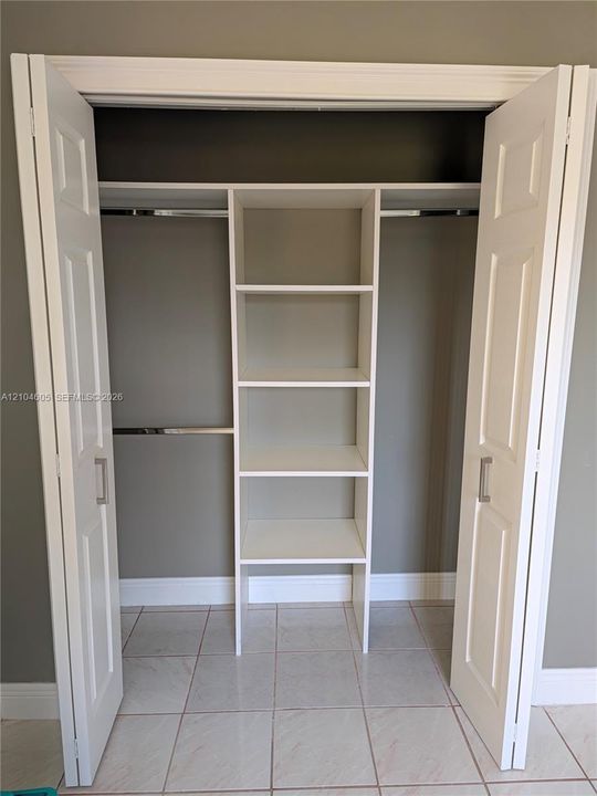 Closet featuring white bi-fold doors, built-in shelving, metal hanging rods, and light-toned tile flooring