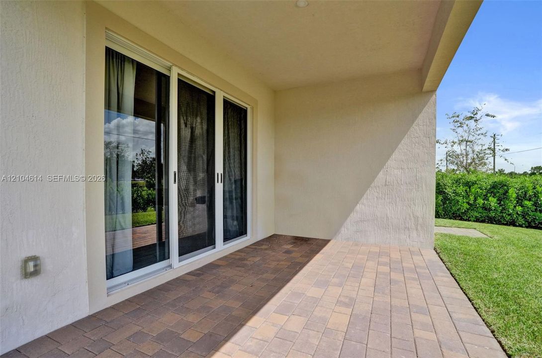Covered patio with multi-tone paver flooring, sliding glass doors, and a surrounding lawn with perimeter hedging