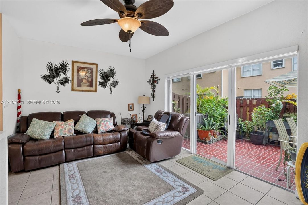 Interior living space featuring large-format ceramic floor tiling and a multi-blade ceiling fan, opening to a brick-paved patio enclosed by a privacy fence