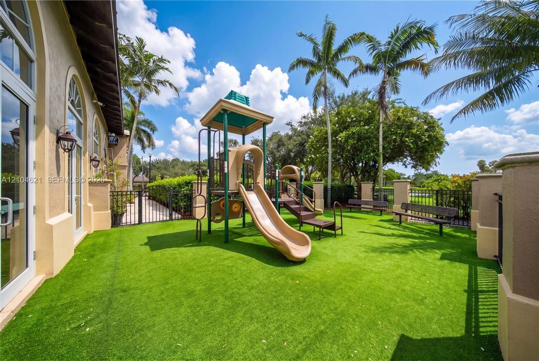 Outdoor recreation area featuring artificial turf, a multi-level playset with integrated slide, metal bench seating, and palm tree landscaping