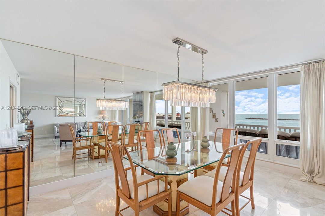 Dining area featuring large-format stone tile flooring, floor-to-ceiling mirror wall, rectangular crystal chandelier, and sliding glass doors with direct water exposure
