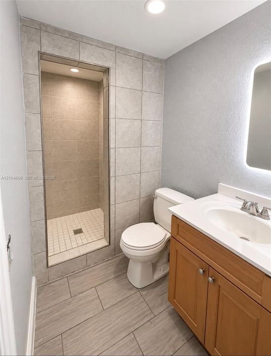 Walk-in shower with floor-to-ceiling tile, vanity with integrated countertop and sink, wood-finish cabinetry, and backlit wall-mounted mirror