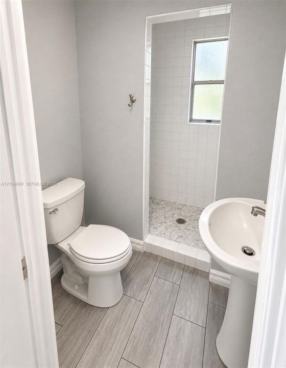 Bathroom featuring wood-finish flooring, a white pedestal sink, a walk-in shower with stone-style pebble flooring, white tile wall accents, and a window