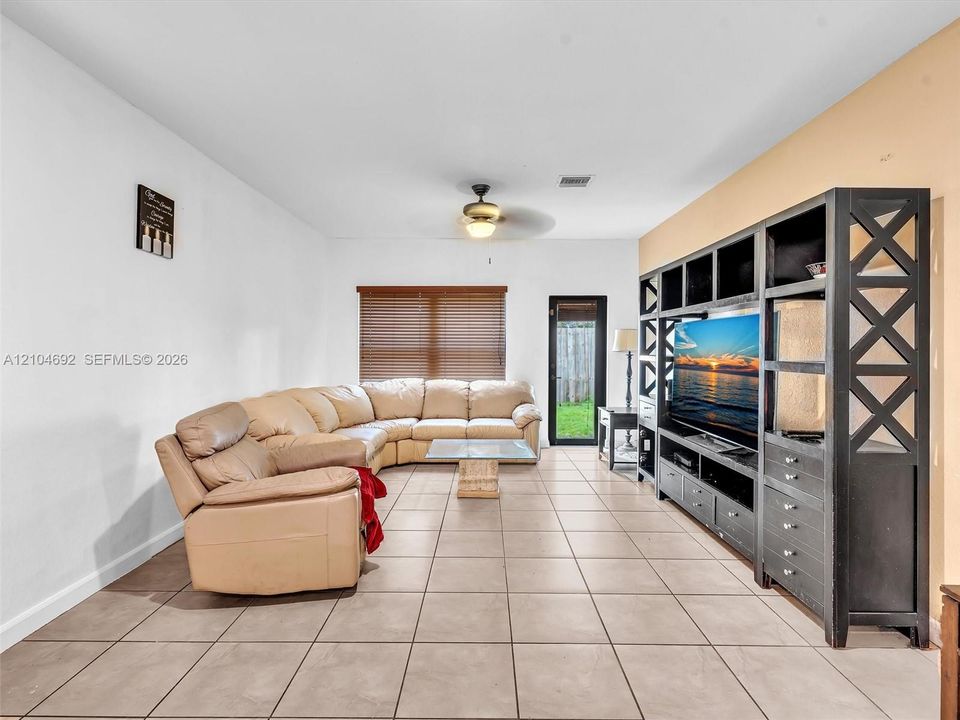 Spacious living area featuring large-format ceramic tile flooring, built-in floor-to-ceiling wooden shelving unit, ceiling fan with integrated lighting, and horizontal window blinds
