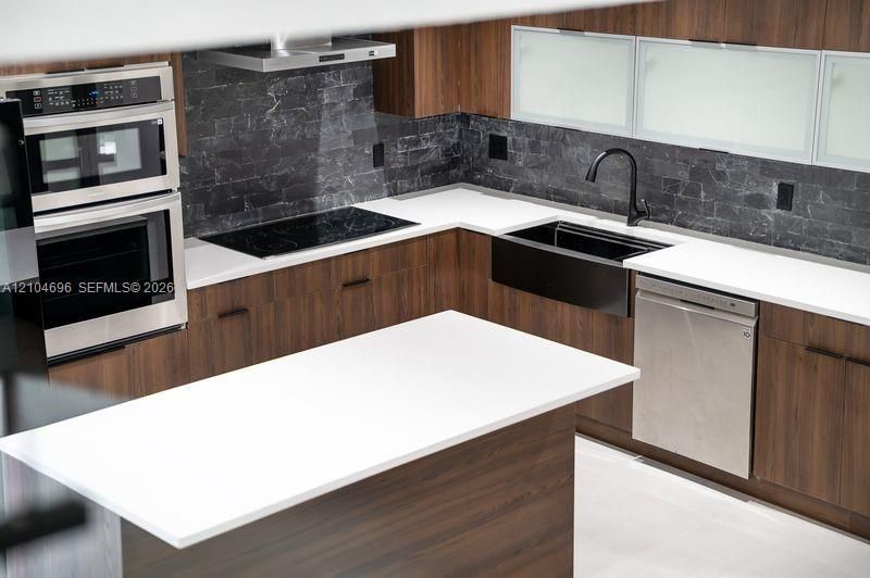 Kitchen featuring wood-finish cabinetry, white countertops, a dark stone backsplash, a black farmhouse-style sink with a matte black faucet, and stainless steel built-in appliances