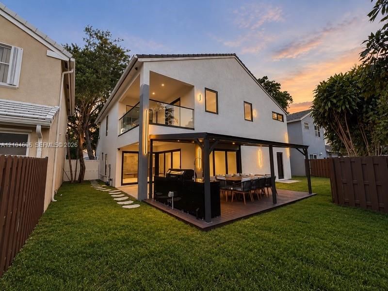 Exterior features include a stucco finish, covered deck with a pergola, built-in outdoor kitchen, second-story balcony with glass railing, and a fenced yard