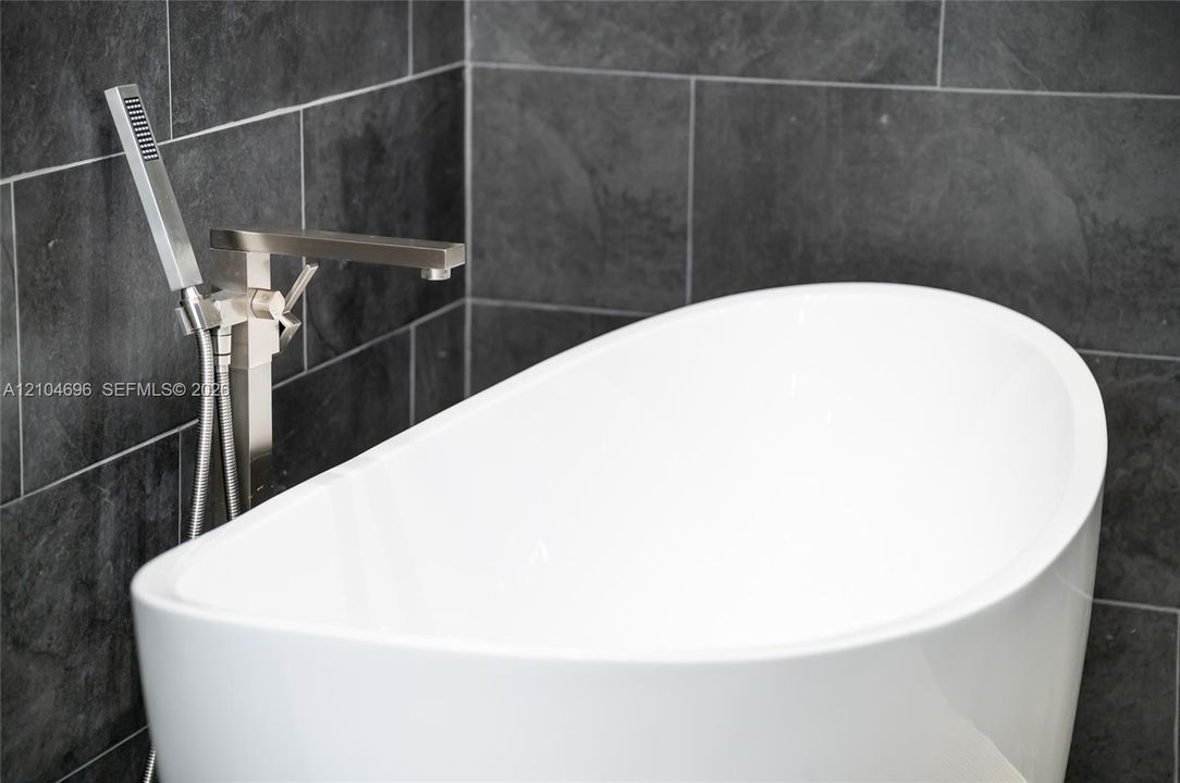 Freestanding white soaking tub featuring a metallic finish floor-mounted faucet with an integrated handheld sprayer, set against charcoal-toned wall tile