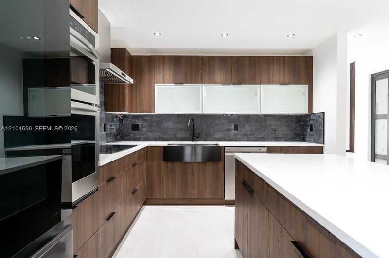 Kitchen featuring wood-finish cabinetry, white countertops, a central island, integrated stainless steel appliances, and a dark stone tile backsplash