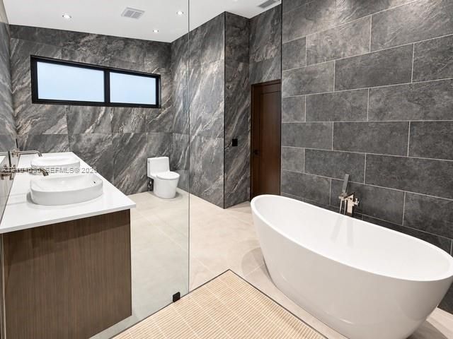 Freestanding white soaking tub with wall-mounted polished chrome faucet, dual vessel sinks atop a wood-finish vanity, and floor-to-ceiling textured stone wall cladding
