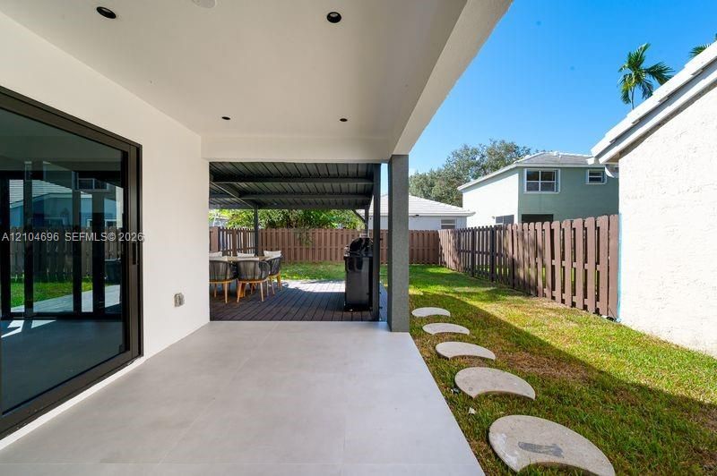 Covered patio featuring large-format porcelain tile flooring and recessed lighting, expanded by a secondary metal-framed outdoor structure with wood-finish decking, complemented by a fenced backyard with stone paver stepping stones