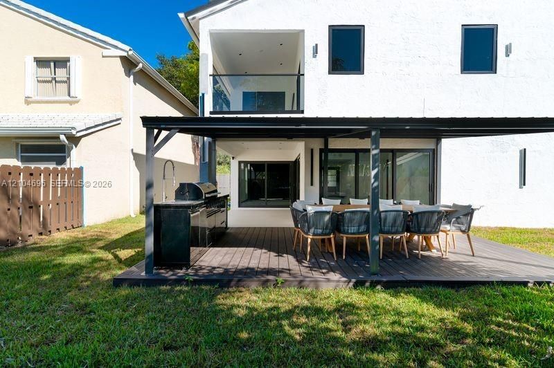 Stucco exterior featuring a covered patio with a dark wood-finish deck, integrated outdoor kitchen, and a glass-railed upper-level balcony