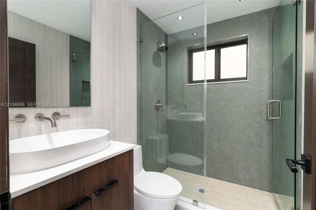 Modern bathroom featuring a wood-finish floating vanity with a white vessel sink, quartz countertop, wall-mounted faucet, and a glass-enclosed shower with textured grey tile surrounds
