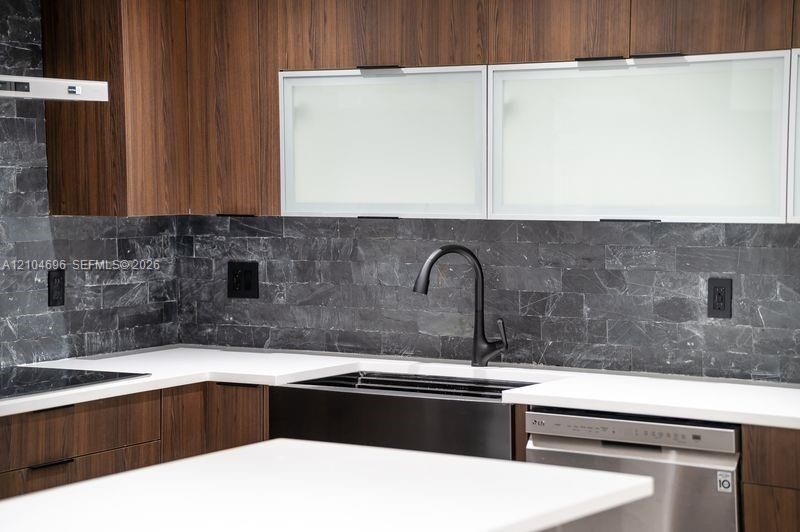 Kitchen featuring wood-finish cabinetry, matte black gooseneck faucet, white solid-surface countertops, textured dark stone backsplash, and frosted glass-panel upper cabinet doors