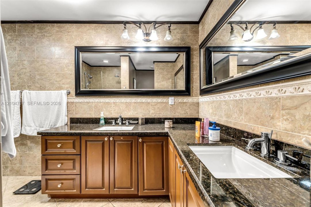 L-shaped wood vanity with stone countertops, dual undermount sinks, full-width wall mirror with dark frame, tiled wall surround with decorative inlay, and multi-bulb vanity light fixture