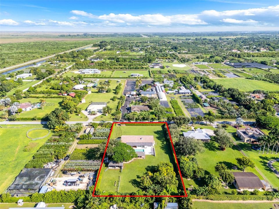 Aerial perspective of a multi-acre property featuring a single-story residence, a fenced-in swimming pool, mature canopy trees, and an expansive lawn