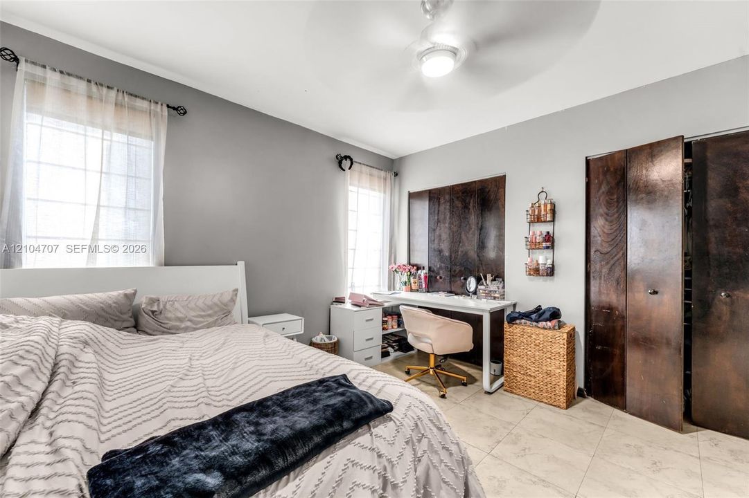 Neutral-toned interior featuring large-format tile flooring, dual-window exposure, a ceiling-mounted light fixture with fan, and bifold closet doors with a wood finish