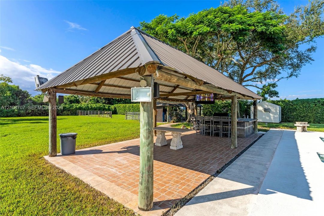 Covered outdoor pavilion featuring a metal roof, log-style timber supports, tile flooring, and a built-in outdoor kitchen island