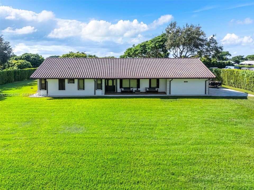 Single-story exterior featuring a clay tile roof, white stucco facade, covered porch, and expansive lawn surrounded by mature hedges