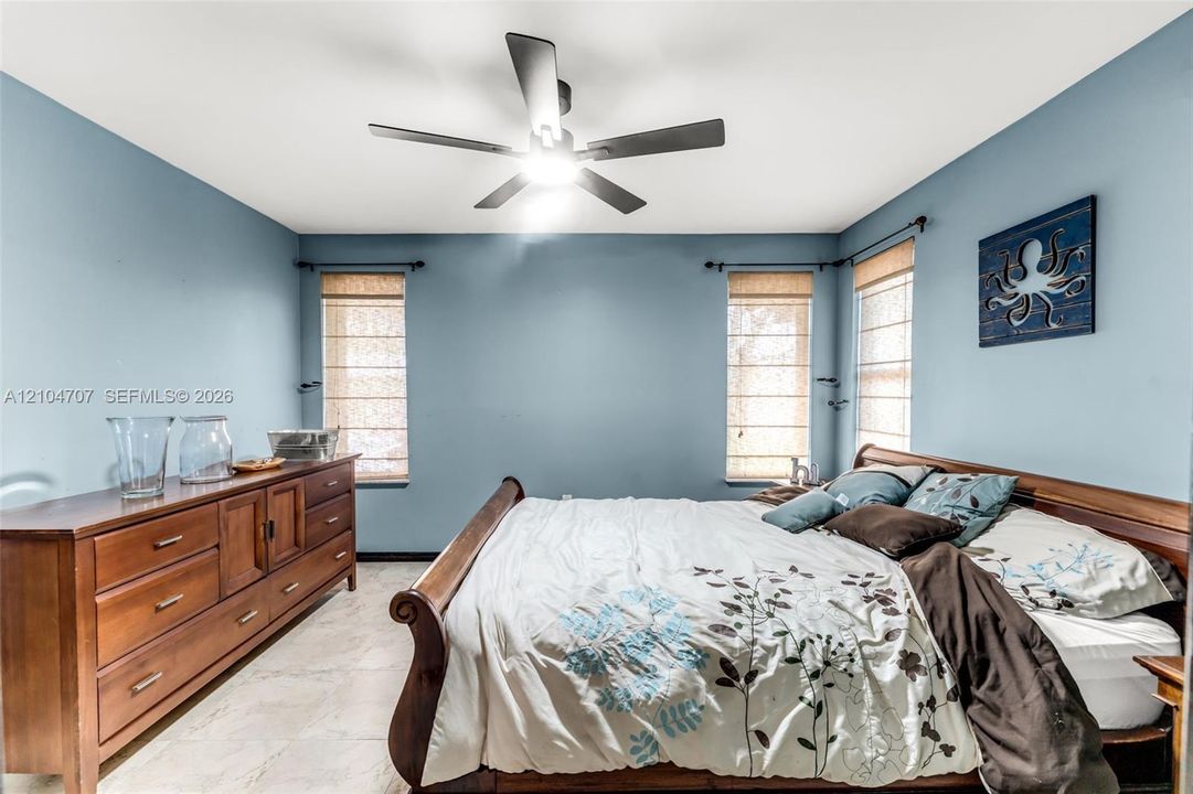 Bedroom featuring tile flooring, neutral wall color, matte black ceiling fan with integrated lighting, and three windows finished with woven-texture Roman shades
