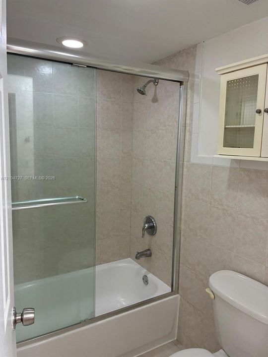 Bathtub and shower combination featuring a sliding glass enclosure, neutral tile surround, recessed lighting, and built-in upper wall cabinetry