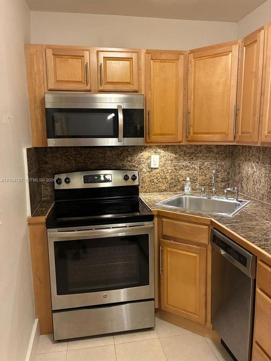 Kitchen featuring light-finish cabinetry, stone-surfaced countertops, tiled backsplash, and a suite of stainless steel appliances including a built-in microwave, range, and dishwasher