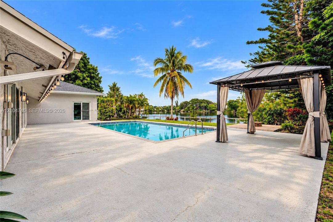 Expansive outdoor living area featuring a rectangular swimming pool, spacious pool deck, and a detached gazebo with privacy drapes, set against a waterfront backdrop