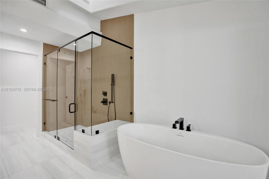 Freestanding soaking tub with matte black fixtures, glass-enclosed shower, shower bench, and light-toned tile flooring