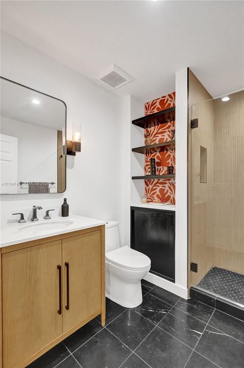 Black tile flooring, single vanity with wood-finish cabinetry, white countertop, modern wall sconce, and a glass-enclosed shower