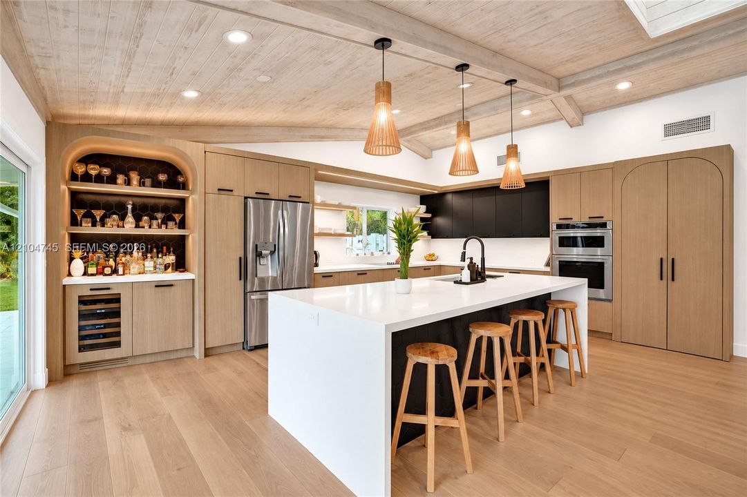 Vaulted wood-finish ceiling with exposed beams, wood-finish flooring, stainless steel appliances, built-in shelving with integrated lighting, and a kitchen island with a white countertop