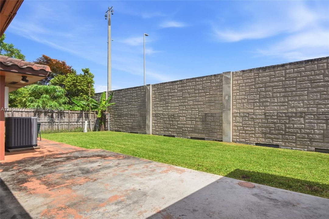 Expansive backyard featuring a manicured lawn, concrete patio, and a tall stone-patterned privacy wall