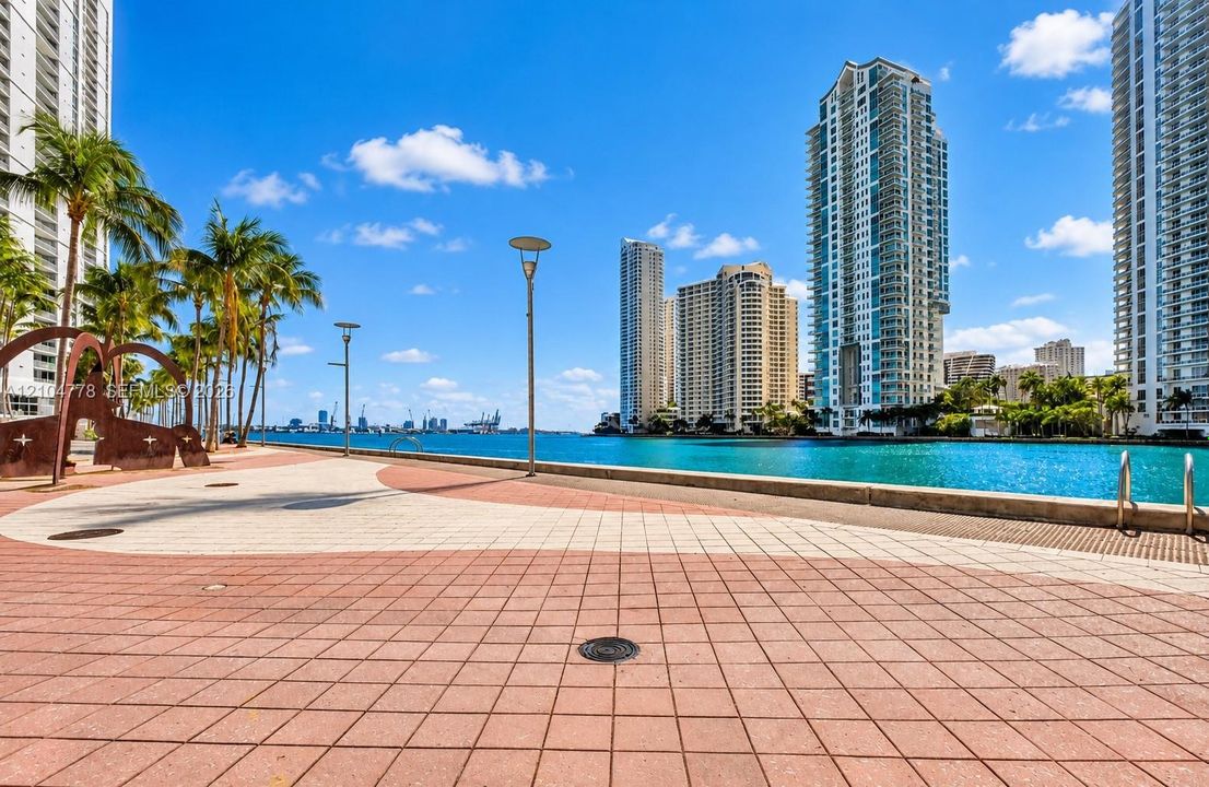 Waterfront promenade featuring a patterned paver walkway, mature palm trees, contemporary streetlights, and a metal art installation