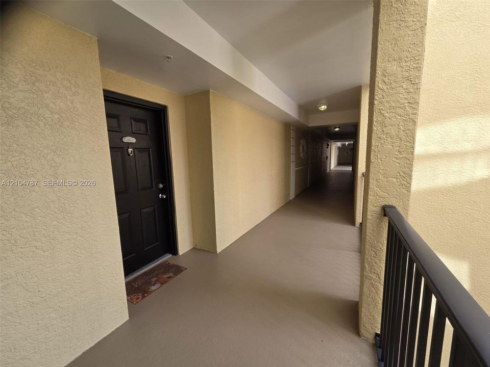 Covered exterior walkway featuring textured stucco walls, black paneled entry door with hardware, flush-mount ceiling lighting, and black metal railings