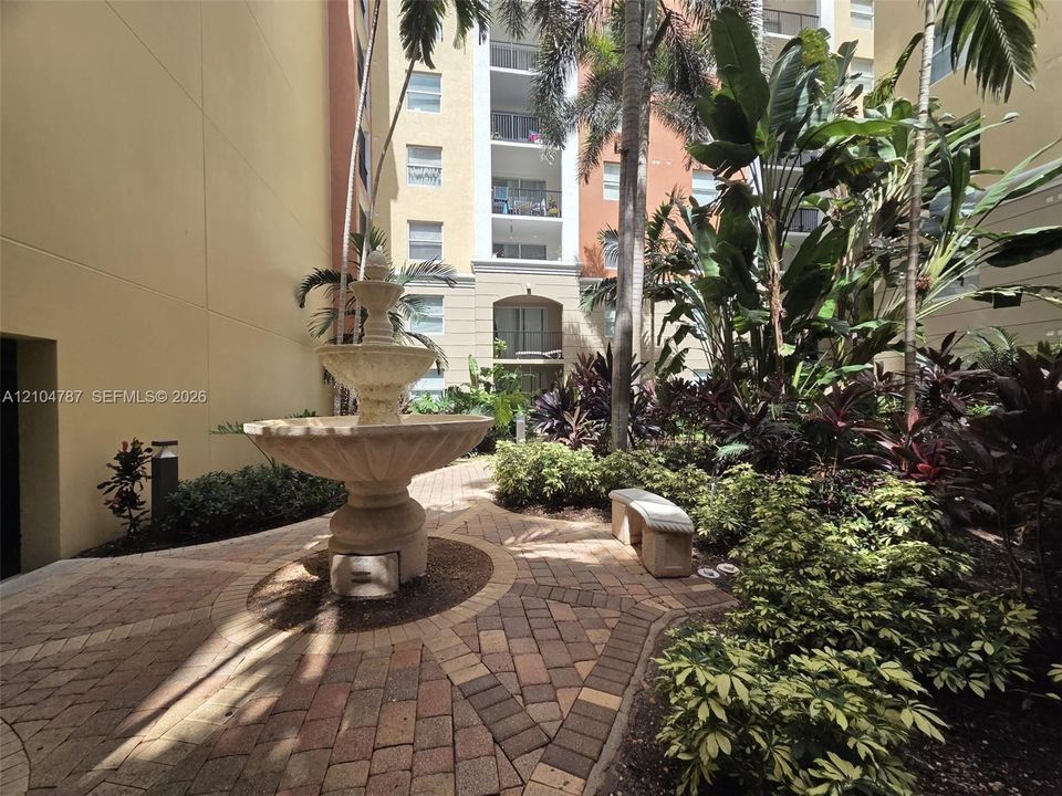 Courtyard featuring a tiered stone fountain, decorative paver hardscaping, mature tropical landscaping, and a permanent stone bench